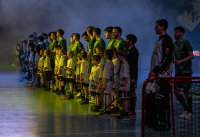 In einer Halle stehen in St. Gallen (9000, Kanton St. Gallen) Spieler und Kinder mit Unihockeyschlaegern gemeinsam aufgereiht vor Spielbeginn unter stimmungsvoller Beleuchtung.