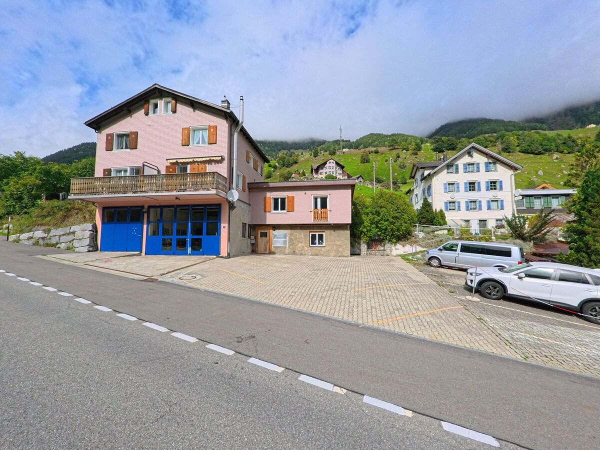 Ein rosa gestrichenes Gebaeude mit blauen Toren steht in Wassen (6484, Kanton Uri) an einer Strasse mit Parkplaetzen und Blick auf die umliegenden Haenge.