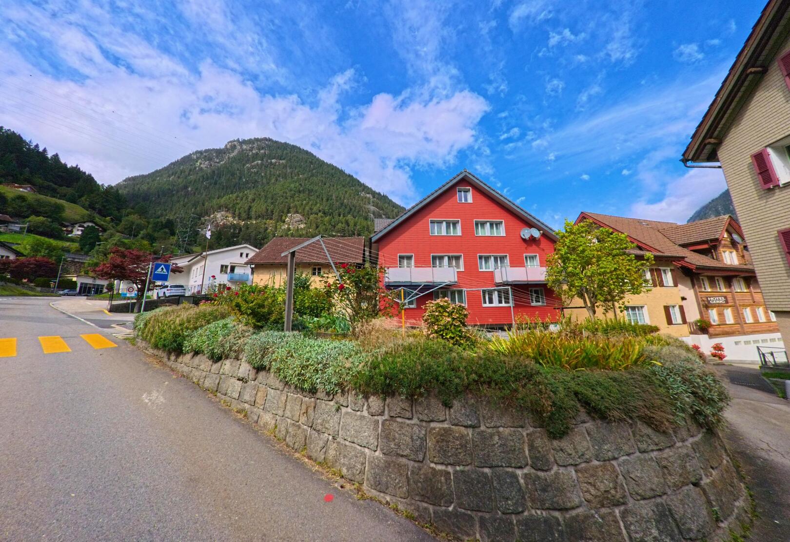 Vor dem roten Mehrfamilienhaus in Wassen (6484, Kanton Uri) stehen Gartenanlagen und im Hintergrund erhebt sich ein bewaldeter Berg.