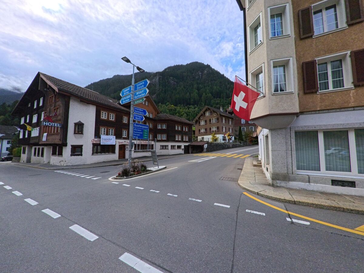 Ein Strassenzug in Wassen (6484, Kanton Uri) zeigt Wegweiser zu verschiedenen Alpenpaessen, daneben haengen an den Gebaeuden Hotel- und Schweizer Fahnen.