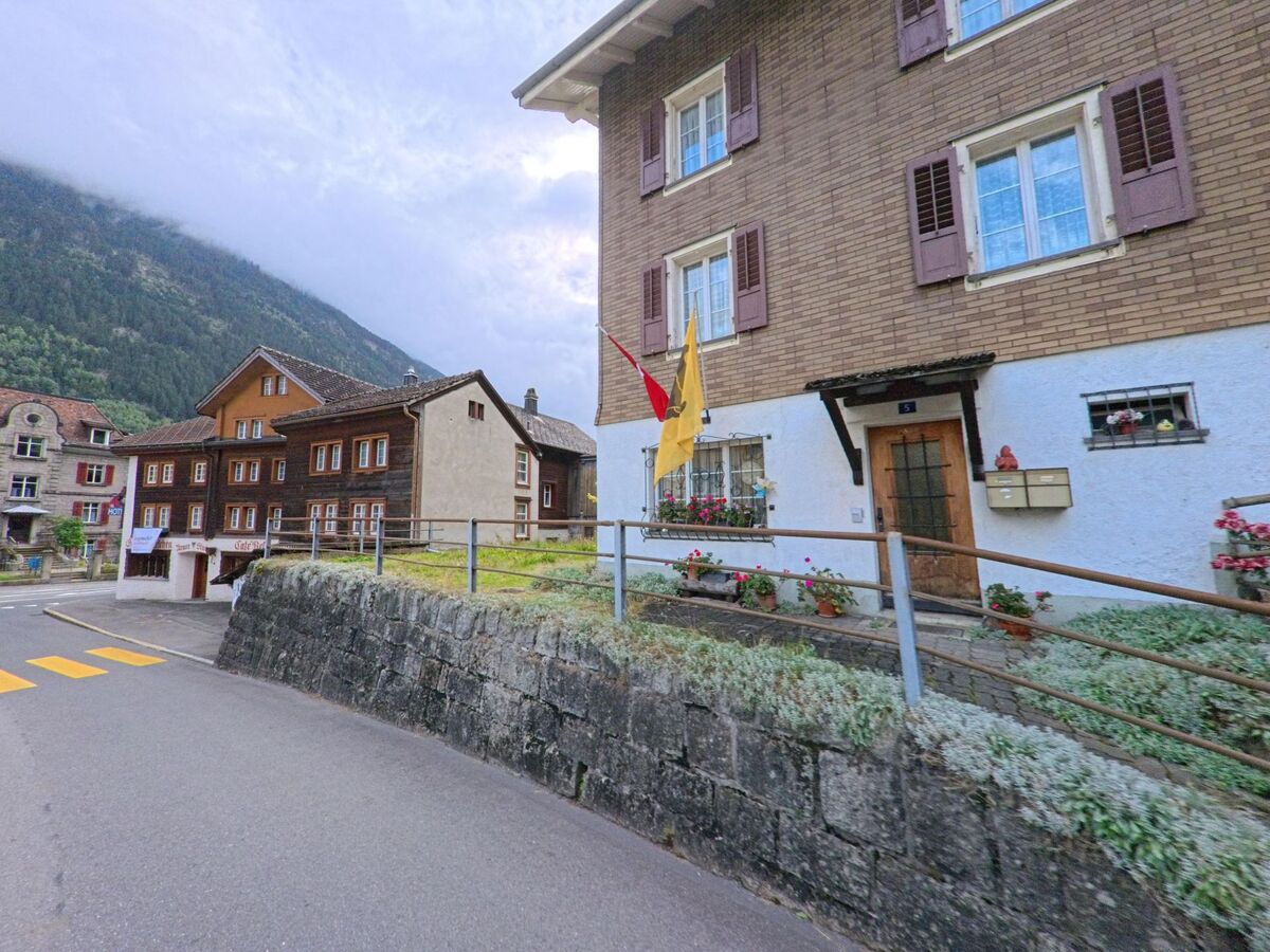 Ein Wohnhaus mit Blumenschmuck und Fahnen steht an einer Strasse in Wassen (6484, Kanton Uri), dahinter sind weitere traditionelle Gebaeude sichtbar.