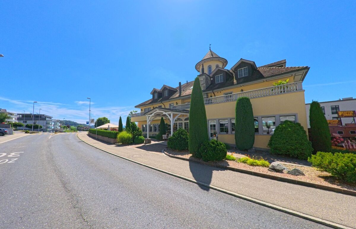 In Wauwil (PLZ 6242, Kanton Luzern) steht ein markantes Gebaeude mit Turm und gepflegter Gartenanlage an einer ruhigen Nebenstrasse bei strahlendem Sonnenschein.