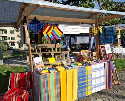 Unter einem Holzstand in Schwarzenburg [3150] im Kanton Bern werden farbige Webarbeiten und Textilien des Webstubenvereins auf einem Markt praesentiert.
