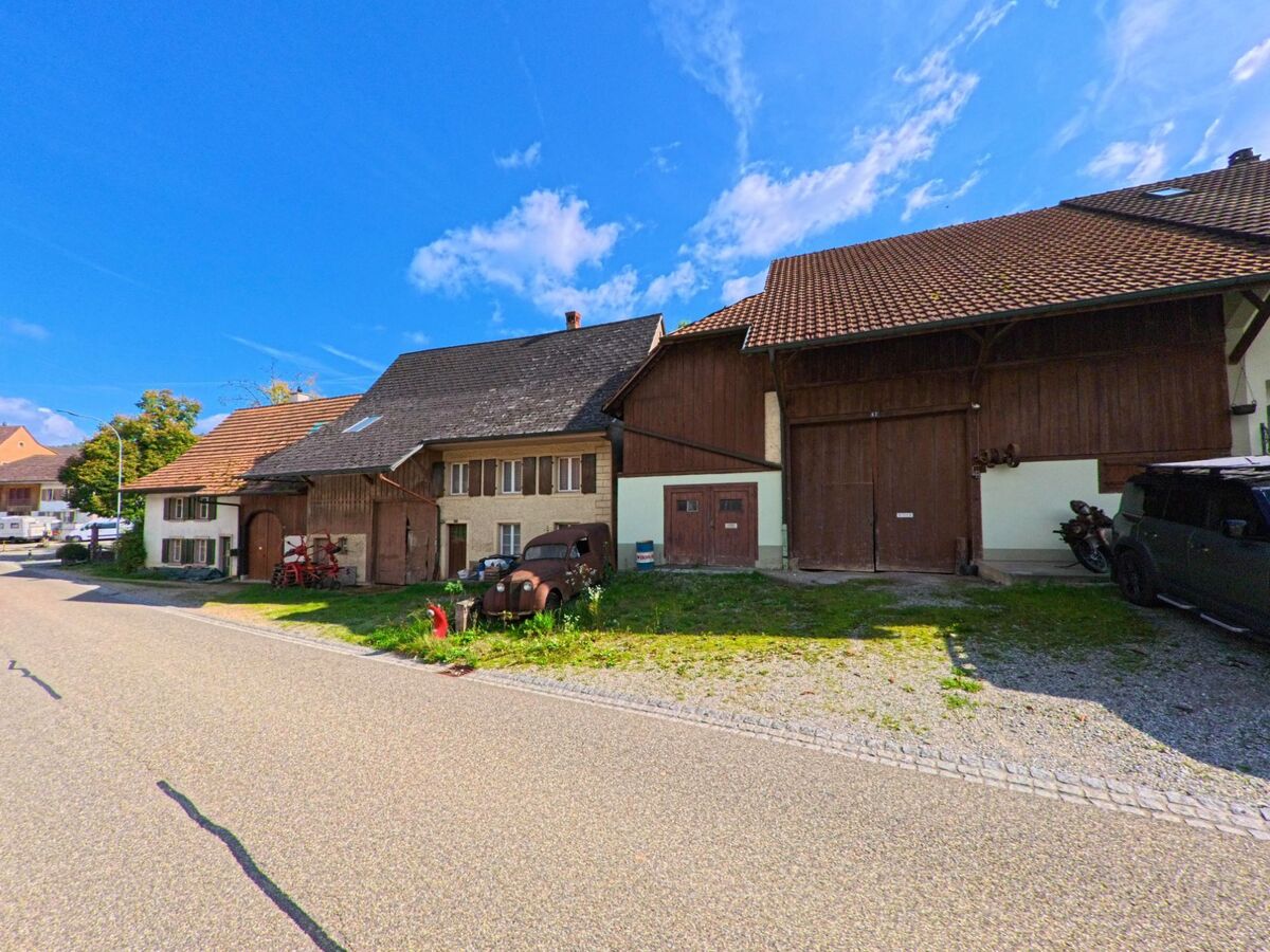 Alte Bauerngebaeude mit Holztoren und verwitterten Details stehen in Wegenstetten (4317, Kanton Aargau) direkt an einer schmalen, sonnigen Strasse.