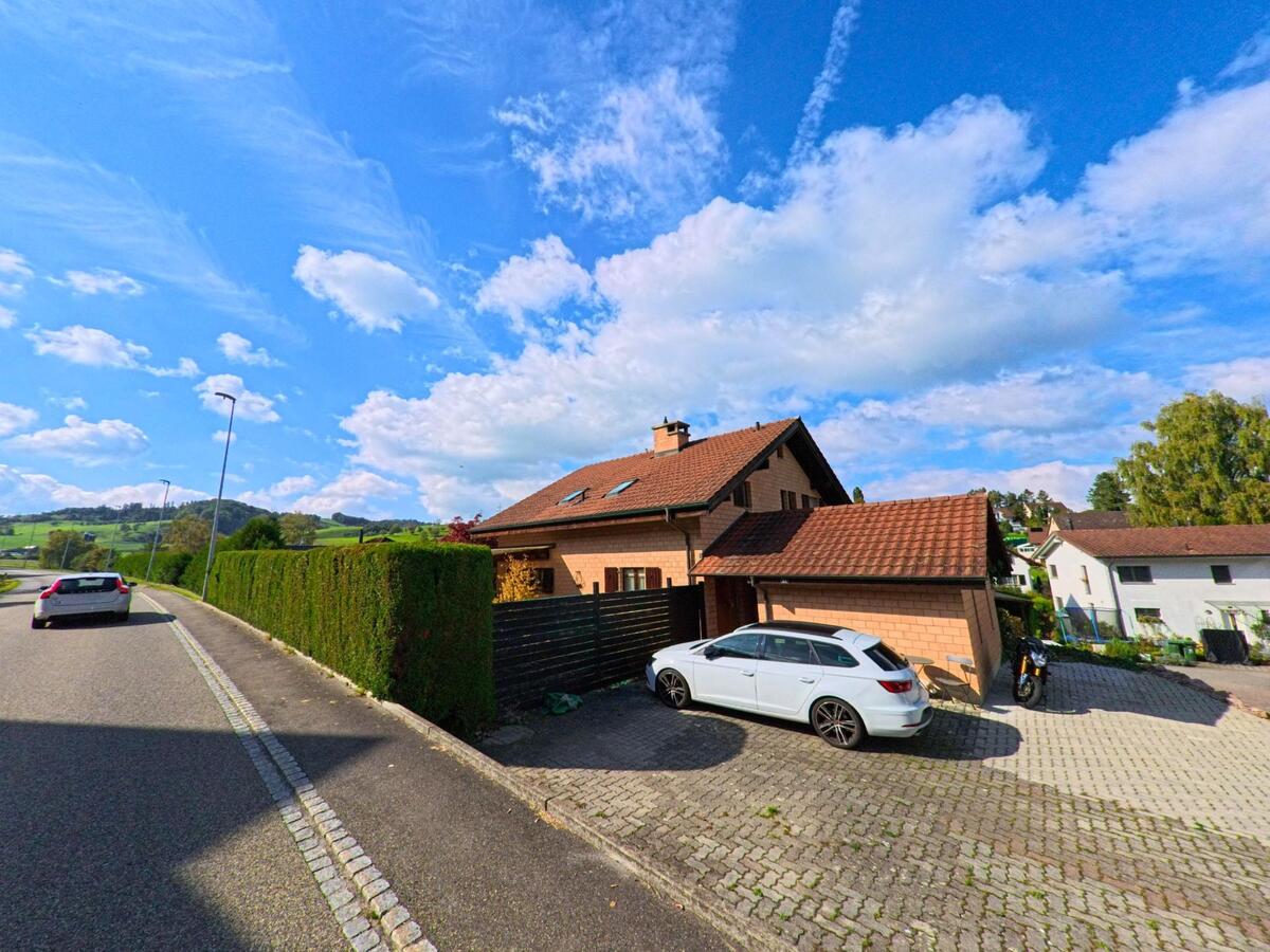 Ein Einfamilienhaus mit grossem Vorplatz liegt in Wegenstetten (4317, Kanton Aargau) am Strassenrand mit Blick auf sanfte Huegel und weite Landschaft.