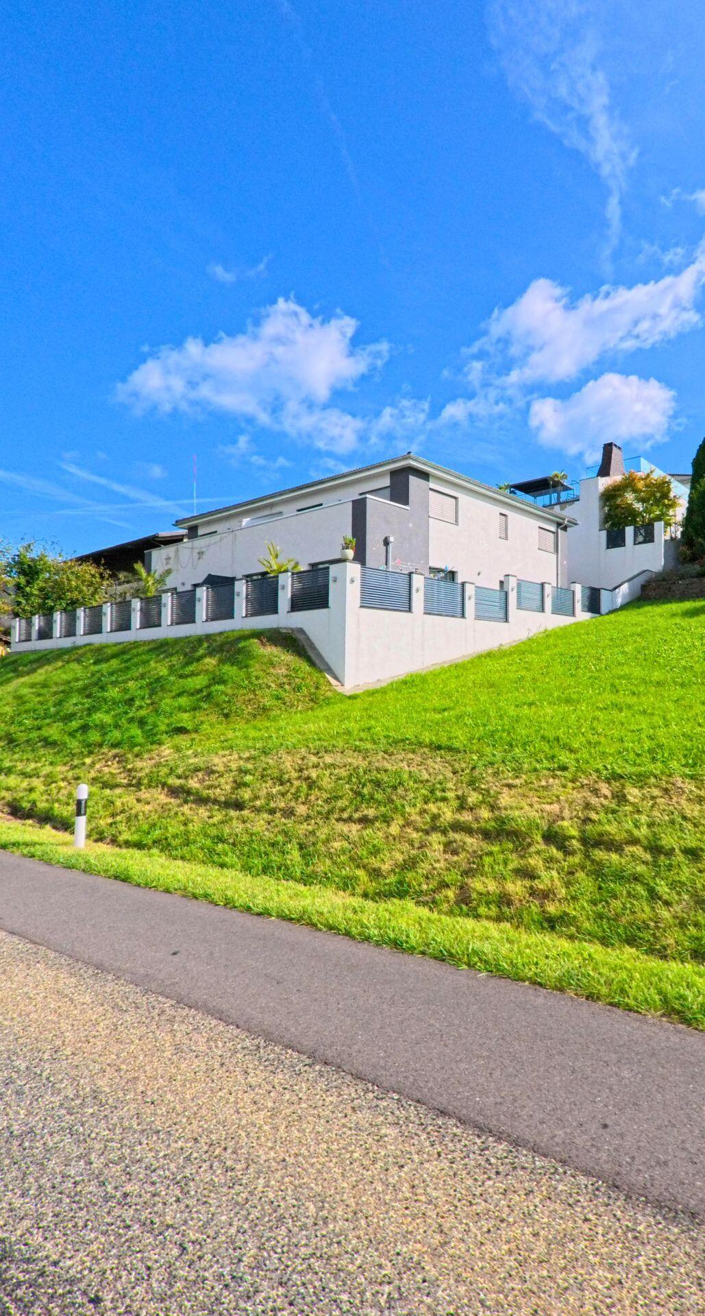 Ein modernes Wohnhaus erhebt sich in Wegenstetten (4317, Kanton Aargau) oberhalb einer steilen, gepflegten Gruenflaeche mit Einfriedung und Terrasse.