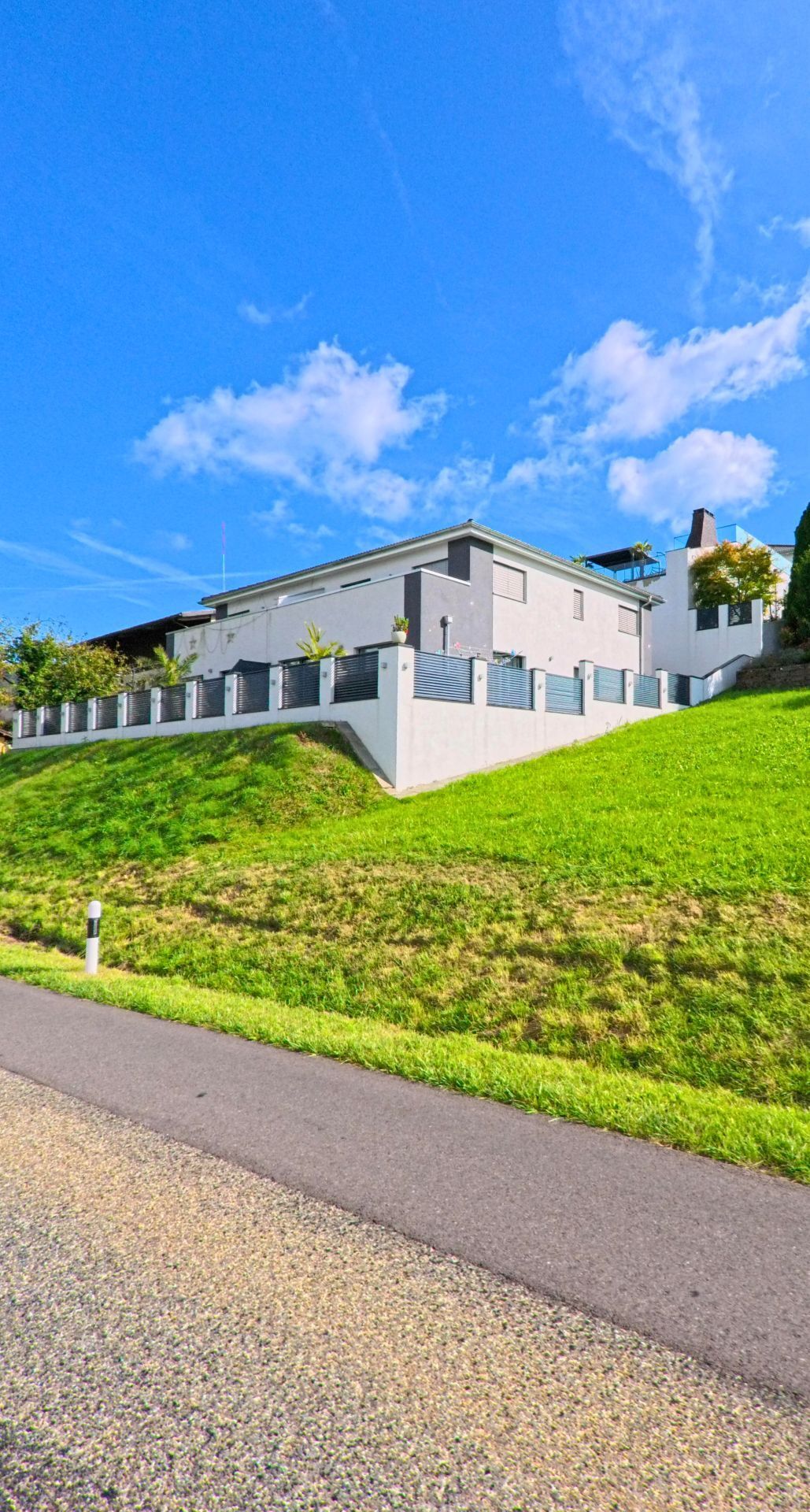 Ein modernes Wohnhaus erhebt sich in Wegenstetten (4317, Kanton Aargau) oberhalb einer steilen, gepflegten Gruenflaeche mit Einfriedung und Terrasse.