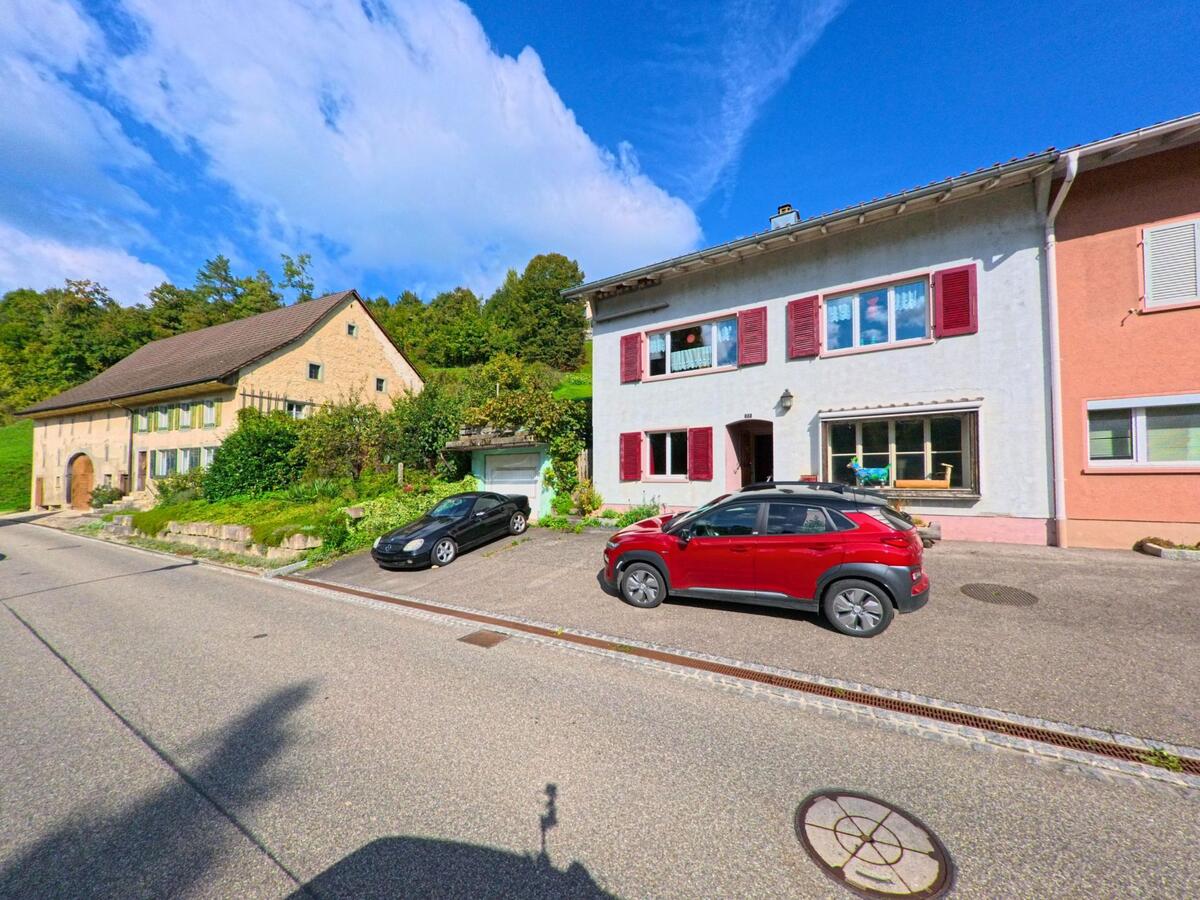 In einer Wohnstrasse von Wegenstetten (4317, Kanton Aargau) stehen ein traditionelles Steinhaus und ein Haus mit roten Fensterlaeden vor bewaldetem Hang.