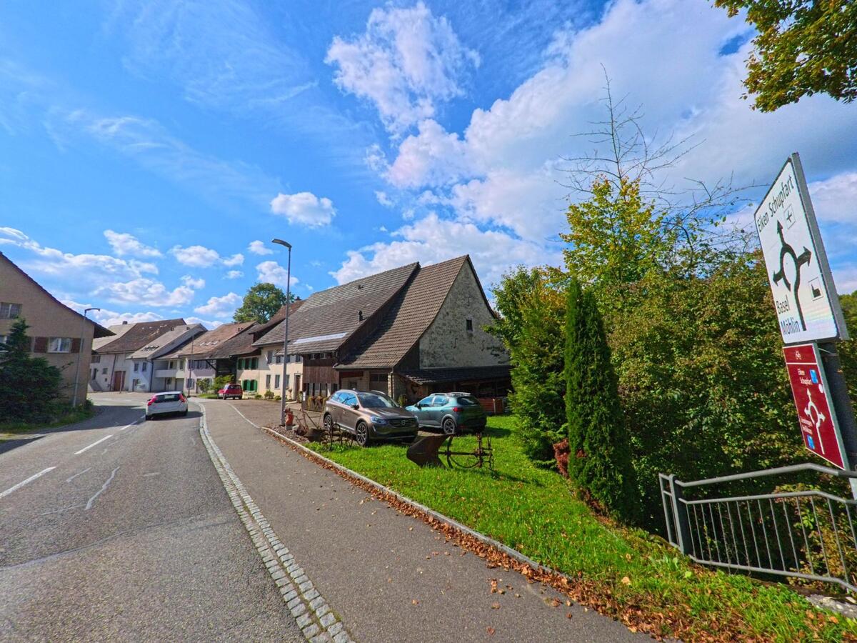In Wegenstetten (4317, Kanton Aargau) fuehrt eine leicht gekruemmte Strasse an traditionellen Wohnhaeusern vorbei, waehrend Wegweiser und Baeume den Rand saeumen.