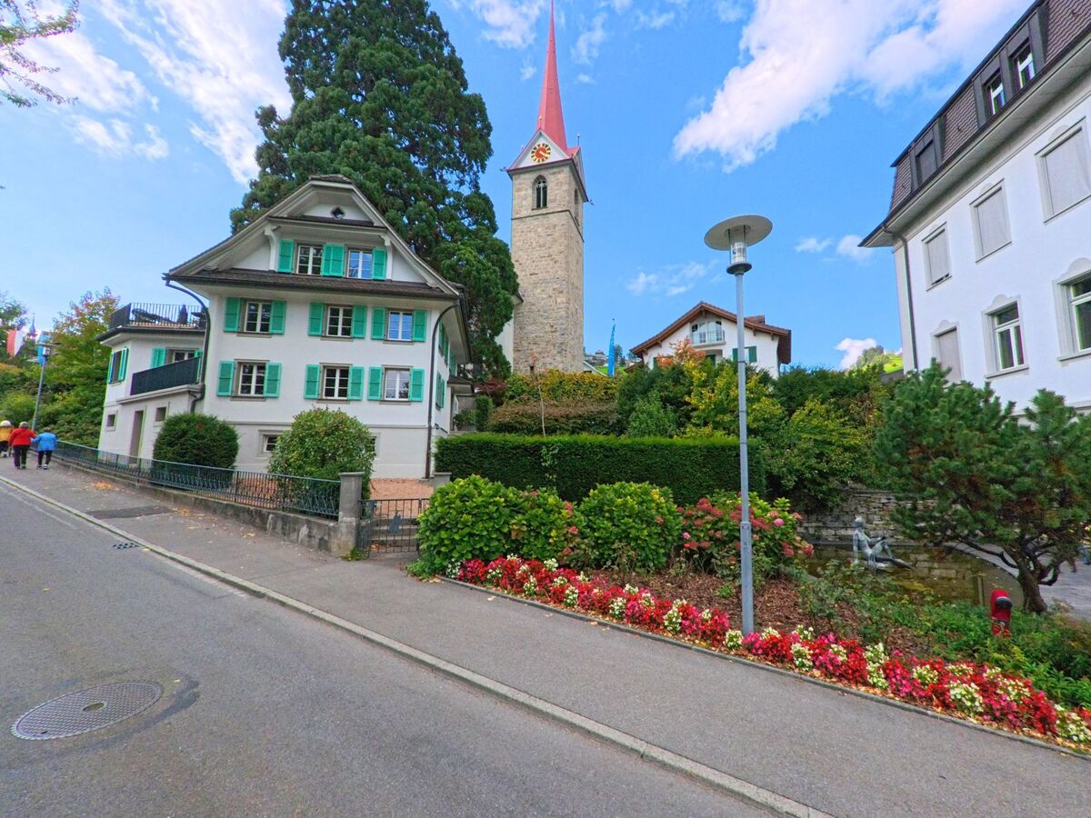 In einer gepflegten Strassenszene in Weggis (6353, Kanton Luzern) stehen ein Haus mit gruenen Fensterlaeden und der Kirchturm zwischen bunten Blumenbeeten und dichter Vegetation.