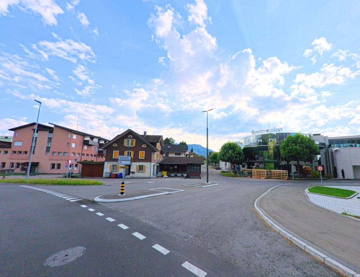 Im Gebiet von Weggis (6353, Kanton Luzern) liegt eine weitlaeufige Kreuzung, umgeben von Wohnhaeusern und einem modernen Firmengebaeude, waehrend sich im Hintergrund Huegel und ein aufgelockertes Wolkenbild zeigen.