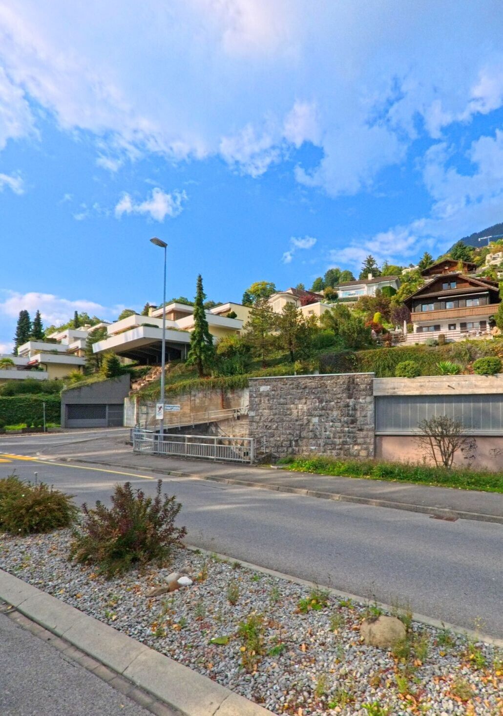 Am Hang von Weggis (6353, Kanton Luzern) stehen moderne und traditionelle Wohnhaeuser zwischen Baumen und Straeuchern, waehrend im Vordergrund eine Strasse mit Verkehrsinsel und niedriger Mauer verlaeuft.