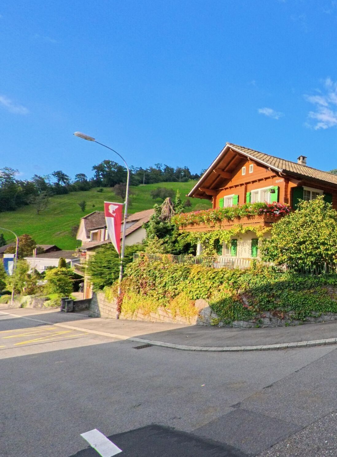 In Weggis (6353, Kanton Luzern) steht ein traditionelles Holzhaus mit gruenen Fensterlaeden und reich bepflanztem Balkon an einer sonnigen Hanglage, waehrend eine Straße und eine Landesflagge den Vordergrund praegen.