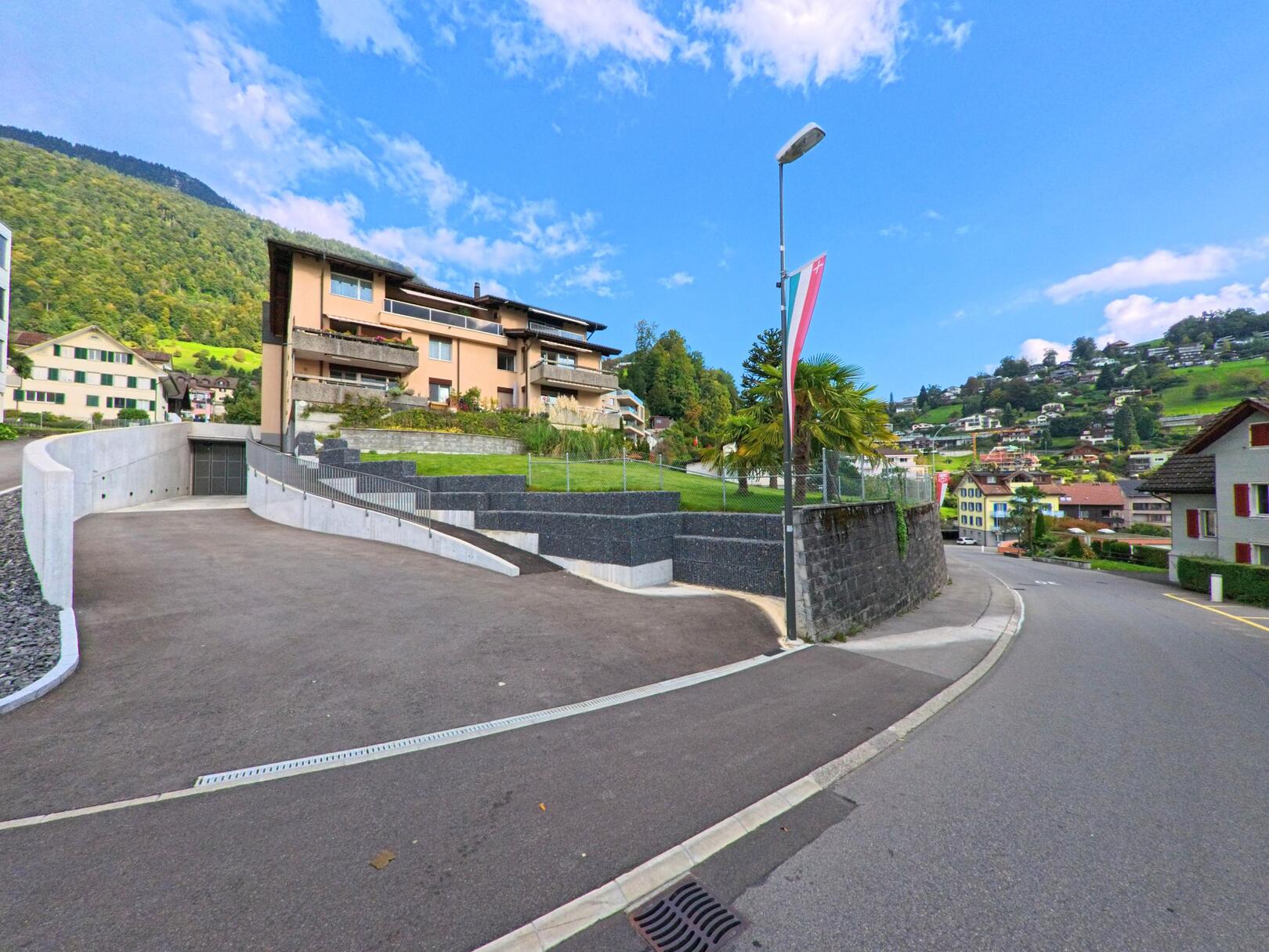 Entlang einer ansteigenden Strasse in Weggis (6353, Kanton Luzern) liegen mehrere Wohnhaeuser vor einem bewaldeten Hang, waehrend eine breite Einfahrt und eine Laterne mit Fahne den Vordergrund bilden.