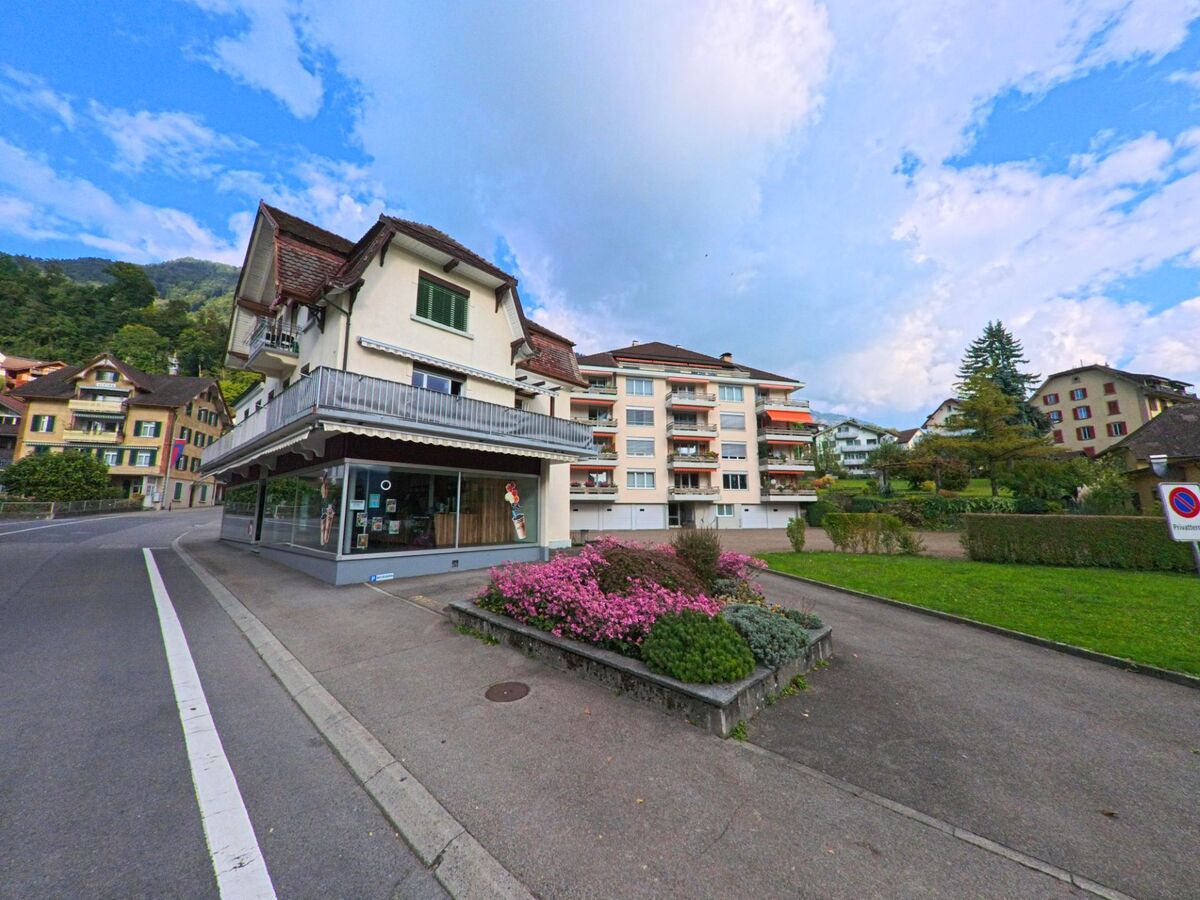 In Weggis (6353, Kanton Luzern) steht ein Wohnhaus mit grossem Balkon an einer Strassenkreuzung, umgeben von weiteren Gebäuden und einem bepflanzten Vorbereich mit rosa Blumen.