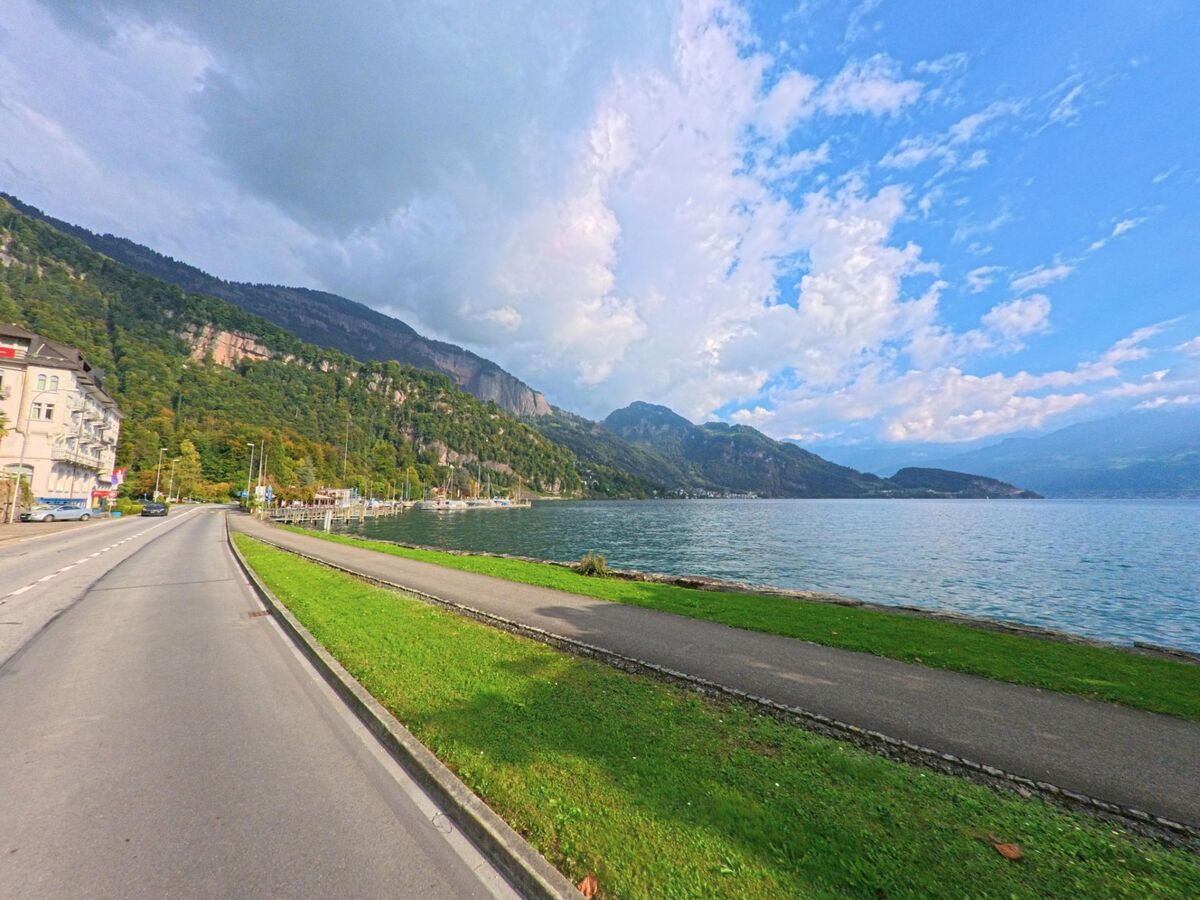 Entlang des Seeufers in Weggis (6356, Kanton Luzern) verlaeuft eine Strasse neben einer Gruenflaeche, waehrend sich rechts der Blick ueber das Wasser auf die umliegenden Berge oeffnet.