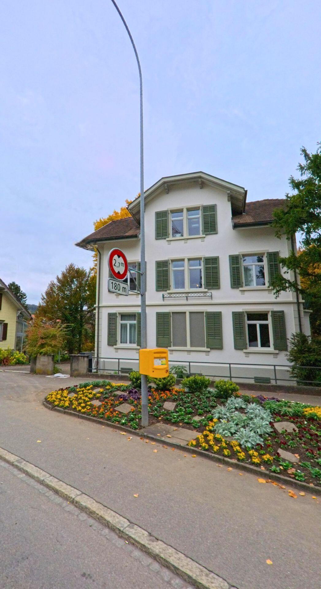 Vor einem gepflegten Blumenbeet steht ein historisches Wohnhaus mit grünen Fensterläden in Weinfelden (8570, Kanton Thurgau) an einer ruhigen Quartierstrasse.