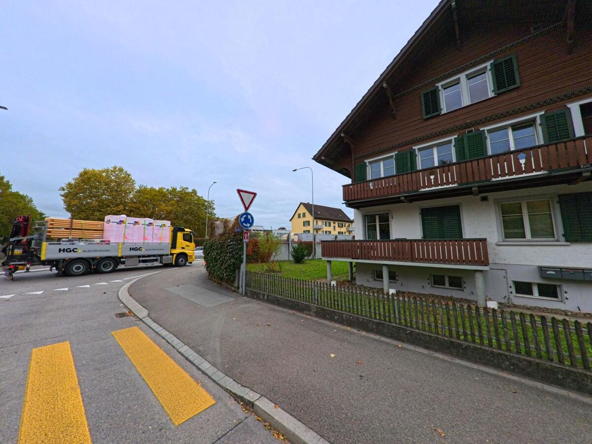 An einer Quartierkreuzung mit Fussgängerstreifen steht ein traditionelles Wohnhaus aus Holz, während ein Lastwagen durch Weinfelden (8570, Kanton Thurgau) fährt.