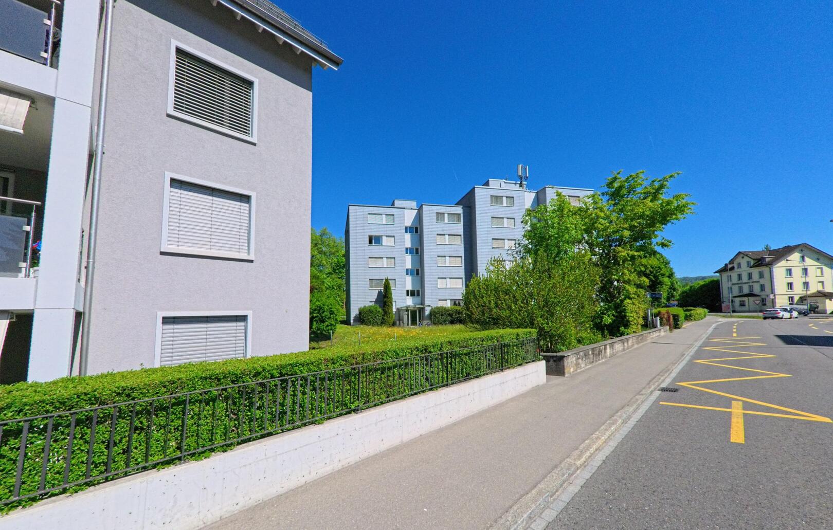 Entlang einer ruhigen Strasse in Weiningen (8951, Kanton Zürich) stehen moderne Mehrfamilienhäuser mit gepflegten Vorgärten und gut sichtbaren Parkmarkierungen.