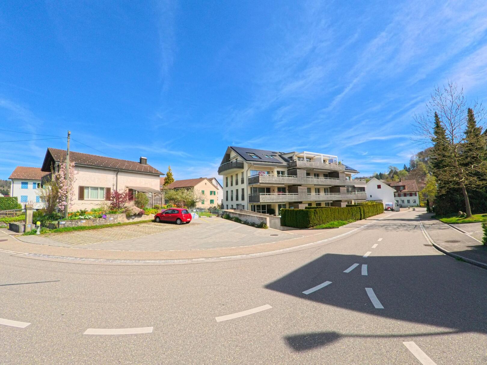 In Weisslingen (8484, Kanton Zürich) führt eine Strasse an modernen Wohnhäusern und einem Parkplatz entlang, auf dem ein rotes Auto geparkt ist, während der Himmel klar und blau ist.