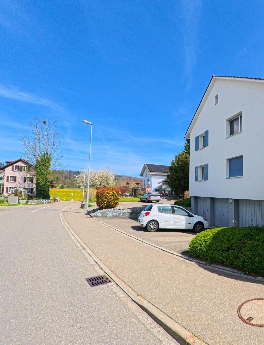 In Weisslingen (8484, Kanton Zürich) führt eine Strasse an modernen Häusern mit Garagen und einem geparkten Auto entlang, während im Hintergrund grüne Wiesen und Hügel zu sehen sind.
