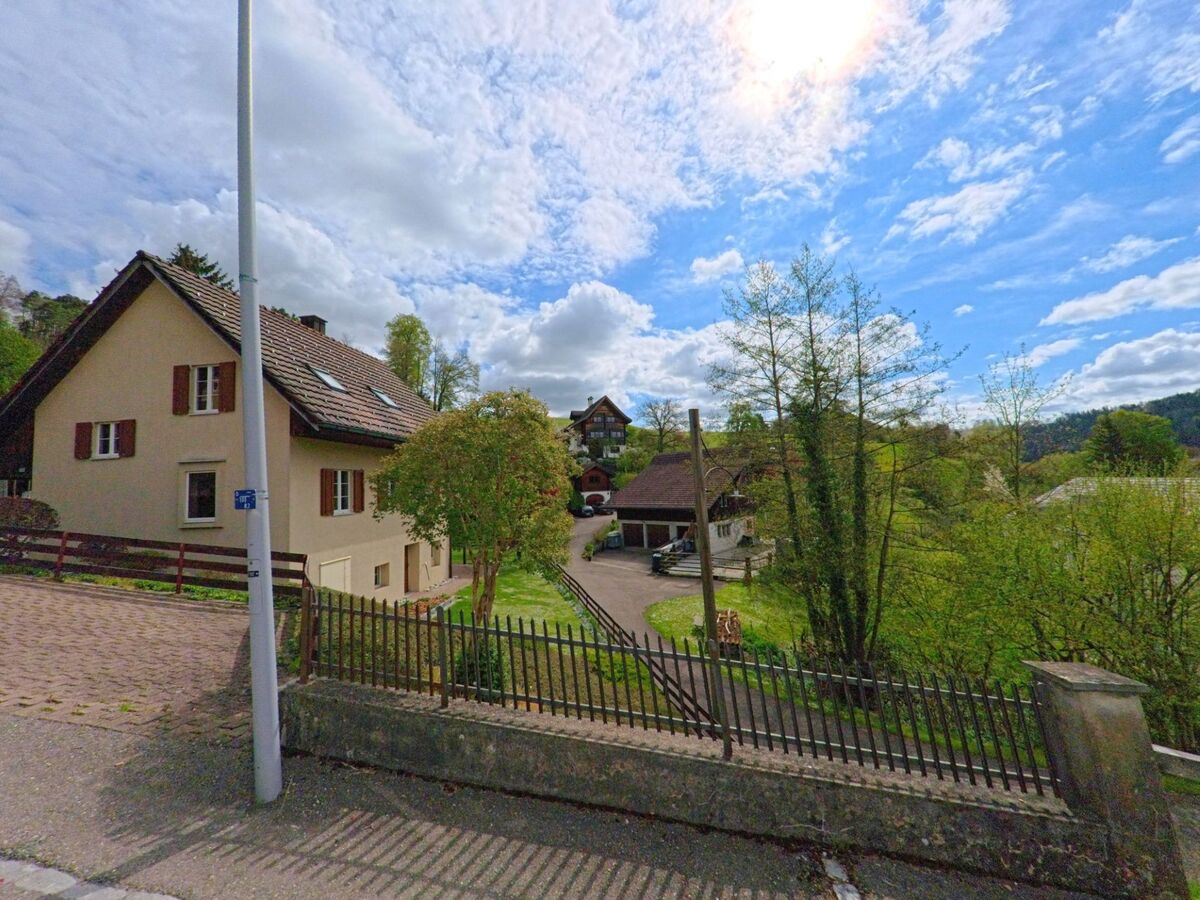 In Weisslingen (8486, Kanton Zürich) führt eine Strasse vorbei an einem traditionellen Haus mit einem gepflegten Garten, während im Hintergrund grüne Hügel und Bäume zu sehen sind.