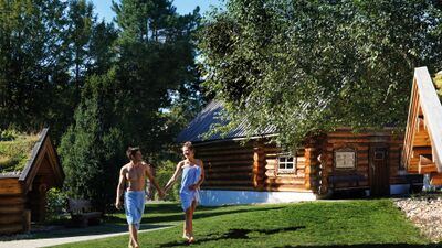 In der Wellness-Welt Sole Uno in Rheinfelden (4310, Kanton Aargau) spaziert ein Paar in Badetuechern durch den grünen Aussenbereich mit rustikalen Blockhuetten.