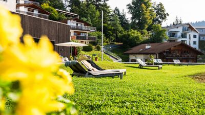 Vor dem WellnessHostel3000 in Laax (7031, Kanton Graubuenden) laden Liegestuehle auf einer gepflegten Wiese mit Blick auf umliegende Hauser und Wald zum Entspannen ein.