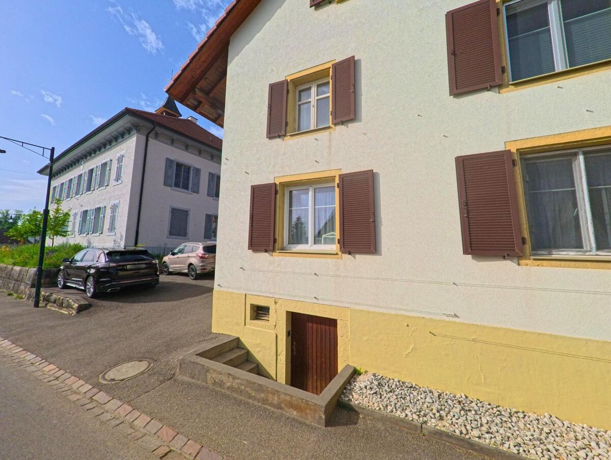 In Wenslingen (4493, Kanton Basel-Landschaft) stehen zwei gepflegte Gebäude mit Fensterläden nahe beieinander, davor parken mehrere Autos an einer kleinen Zufahrt.