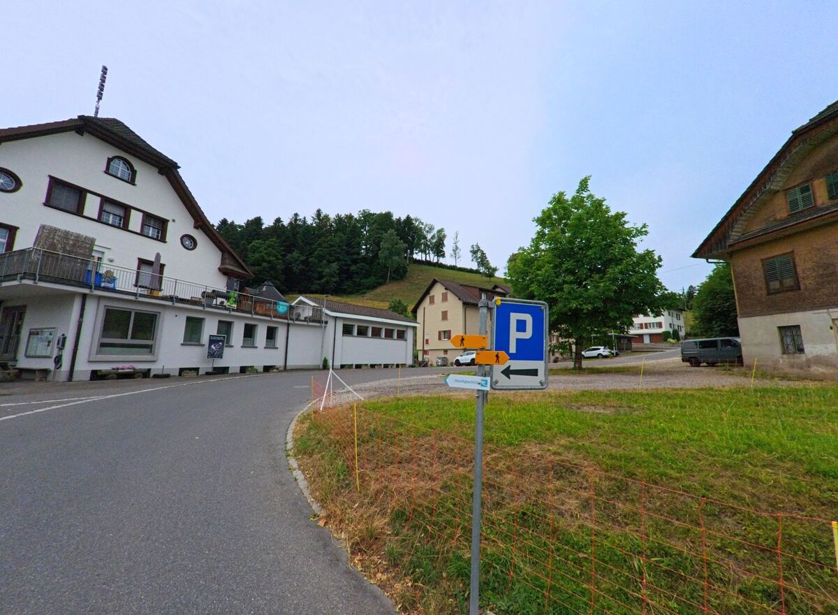 In Werthenstein (6106, Kanton Luzern) zweigt eine Nebenstrasse bei einem Parkplatzschild ab, gesäumt von Wohnhäusern, einem Wegweiser zur Bushaltestelle und einem Baum auf einer Wiese.