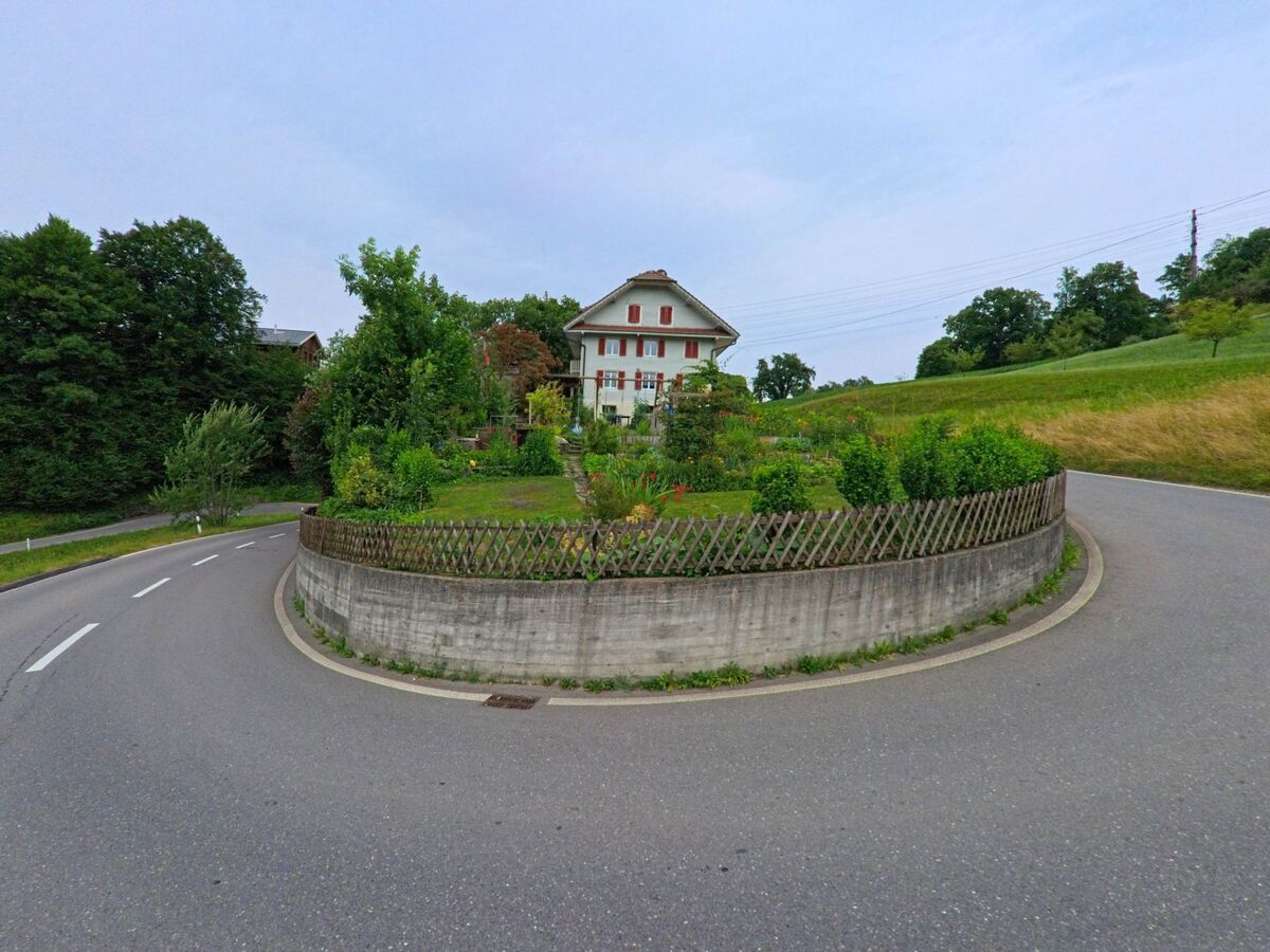 In einer scharfen Kurve bei Werthenstein (6106, Kanton Luzern) steht ein gepflegtes Wohnhaus mit rotem Fensterladen und vorgelagertem Garten, umgeben von Wiesen und Wald.