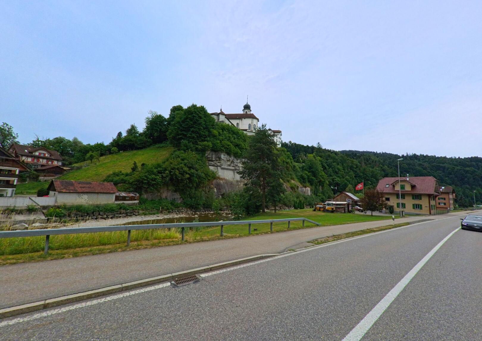 In Werthenstein (6106, Kanton Luzern) thront das Kloster oberhalb einer Felswand über dem Tal, während im Vordergrund eine Landstrasse und verstreute Wohnhäuser zu sehen sind.