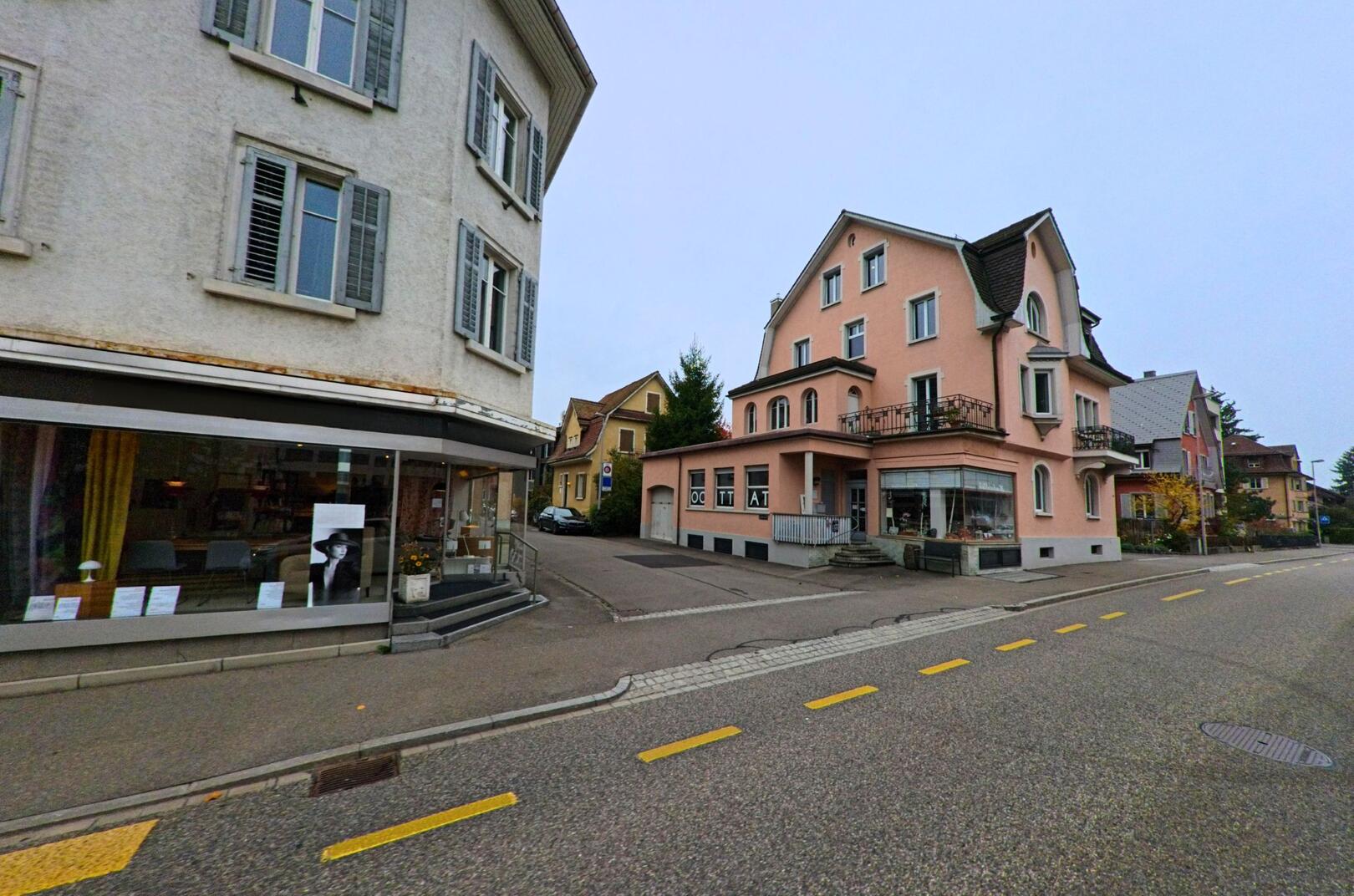Zwischen Wohnhaeusern in Wettingen (5430, Kanton Aargau) verlaeuft eine ruhige Strasse mit kleinen Geschaeften und gedämpftem Tageslicht.