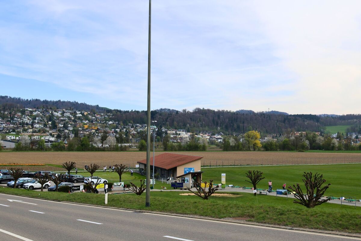 In Wettswil am Albis (8907, Kanton Zürich) liegt eine Golfanlage mit kleinem Clubhaus und Parkplatz am Rande eines grossen Feldes, vor der Kulisse eines bewaldeten Hangs mit Wohnsiedlungen.