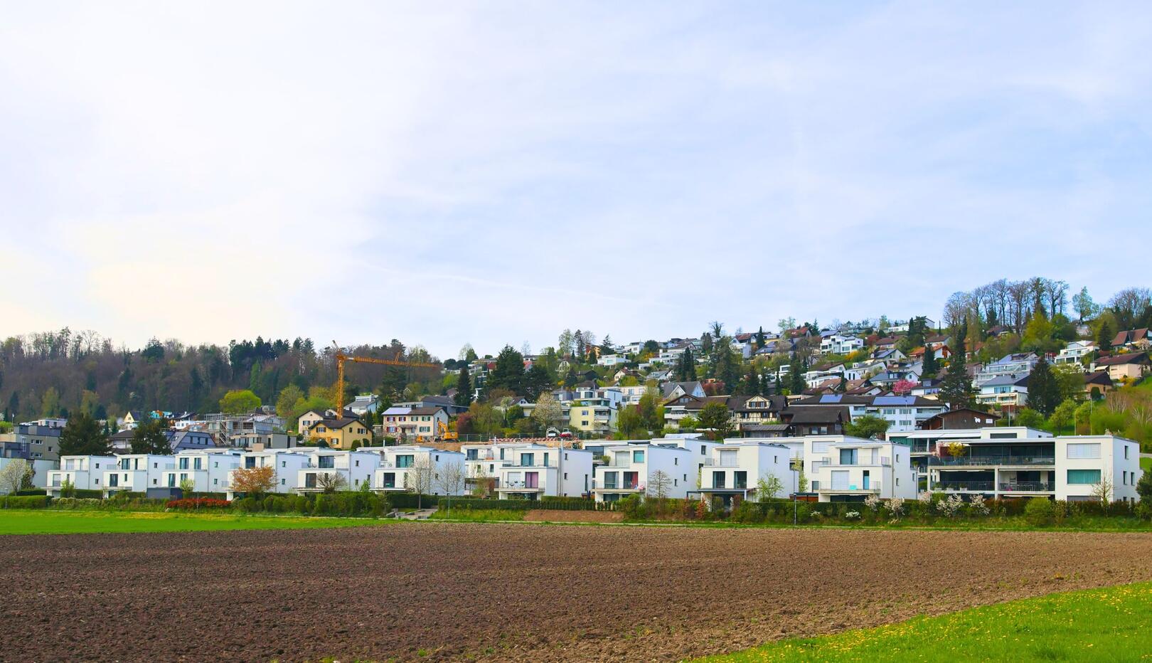 In Wettswil am Albis (8907, Kanton Zürich) erstreckt sich eine moderne Wohnsiedlung entlang eines Hügels, mit einem fruchtbaren Feld im Vordergrund und einem Kran, der auf der Baustelle im Hintergrund sichtbar ist.