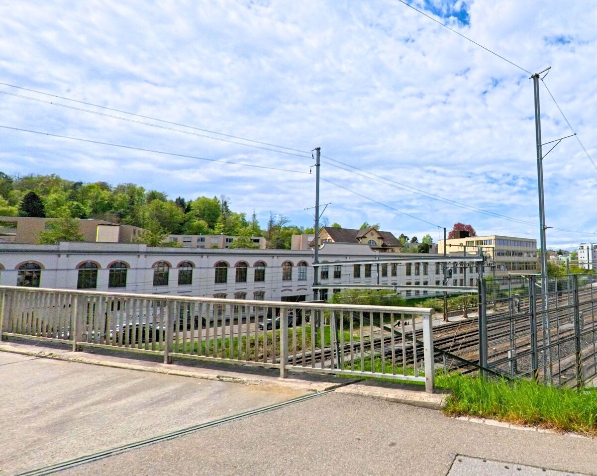 In Wetzikon (8620, Kanton Zürich) überquert ein Gehweg die Bahngleise, flankiert von Werbetafeln und mit Blick auf Industrie- und Verwaltungsgebäude.