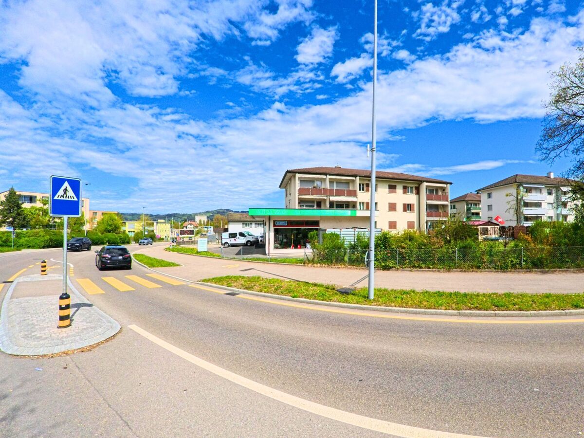 In der Gemeinde Wetzikon (8620, Kanton Zürich) verläuft eine breite Strasse mit Zebrastreifen entlang eines Geschäftshauses und mehrstöckiger Wohnbauten.