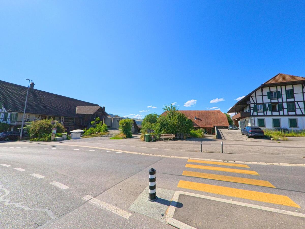  In Wichtrach (3114, Kanton Bern) stehen zwei traditionelle Bauernhäuser mit Fachwerkfassaden an einer Kreuzung mit Zebrastreifen und breitem Vorplatz.