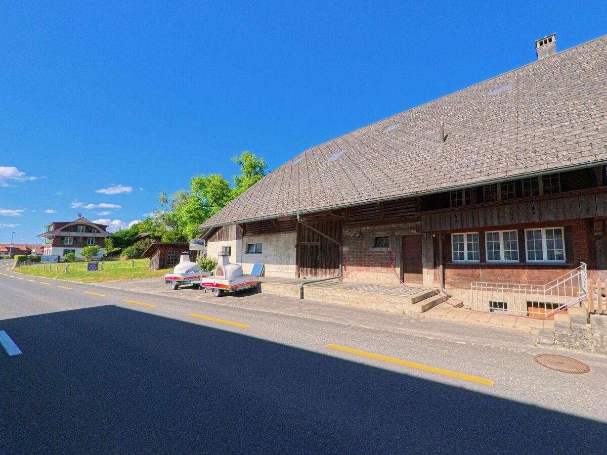 In Wichtrach (3114, Kanton Bern) erstreckt sich ein grosses Bauernhaus mit tief gezogenem Walmdach entlang einer sanft geschwungenen Strasse, vor dem zwei Anhänger geparkt sind.