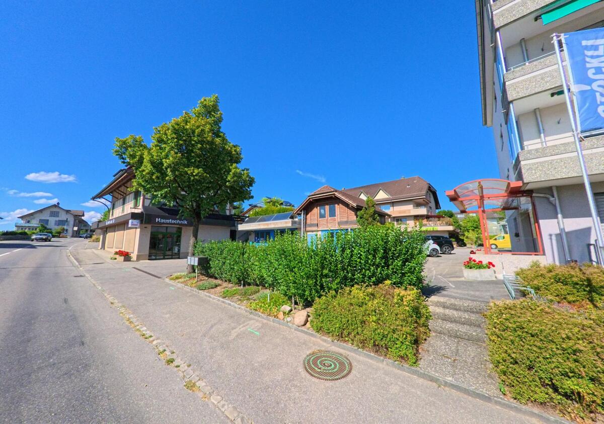 In Wichtrach (3114, Kanton Bern) befinden sich entlang einer leicht ansteigenden Strasse Wohn- und Geschäftshäuser, darunter ein Haustechnikladen und ein Mehrfamilienhaus mit markanter Eingangsüberdachung.