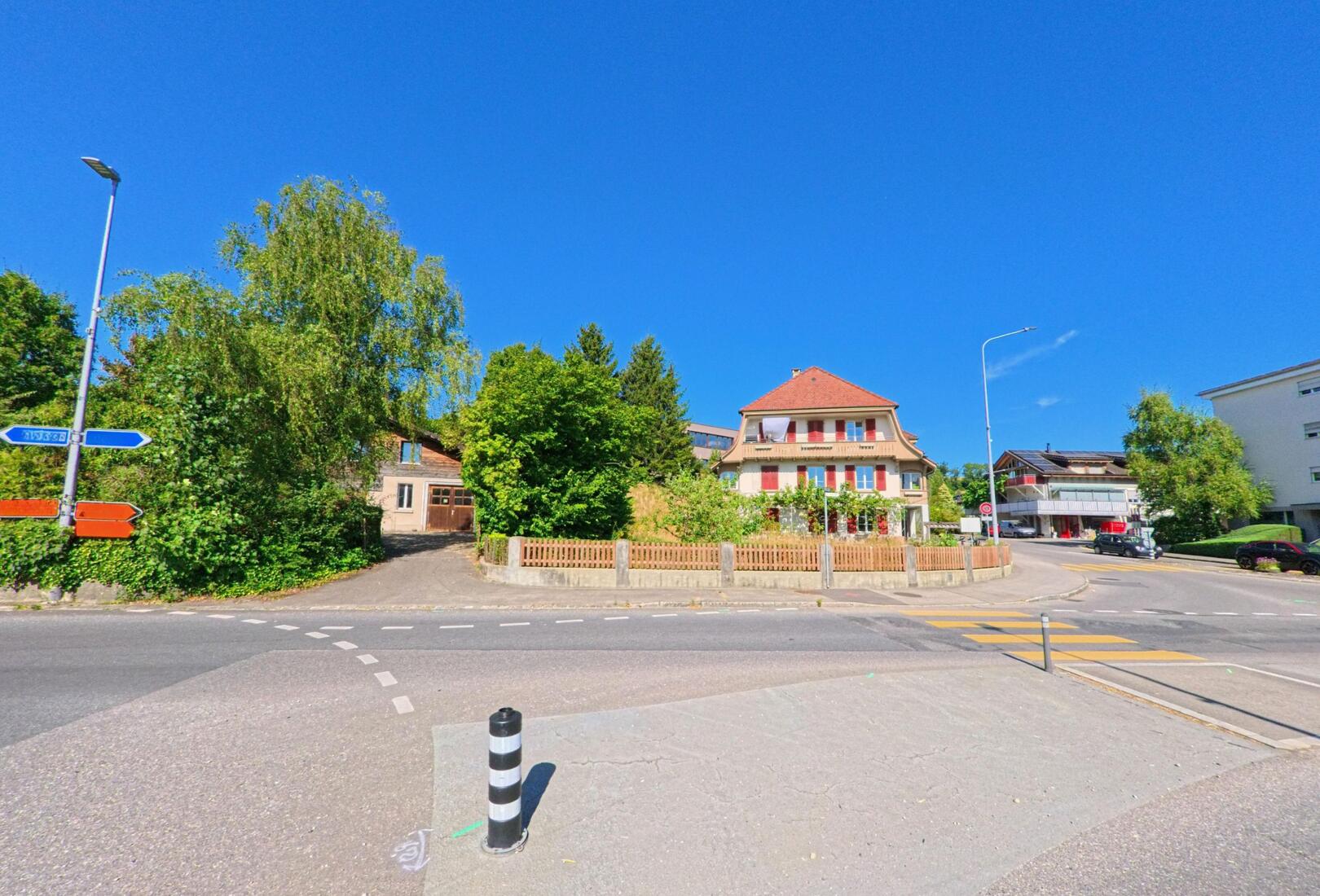 In Wichtrach (3114, Kanton Bern) steht ein Wohnhaus mit roten Fensterläden an einer Kreuzung, wo Wegweiser die Fahrtrichtung nach Bern und Thun anzeigen.