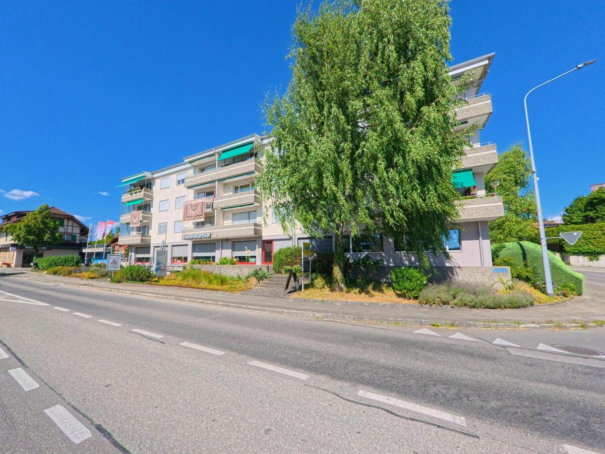 In Wichtrach (3114, Kanton Bern) führt eine kurvige Strasse an einem Wohnhaus mit roten Fensterläden und gepflegtem Vorgarten vorbei, während im Hintergrund ein Motorroller vorbeifährt.