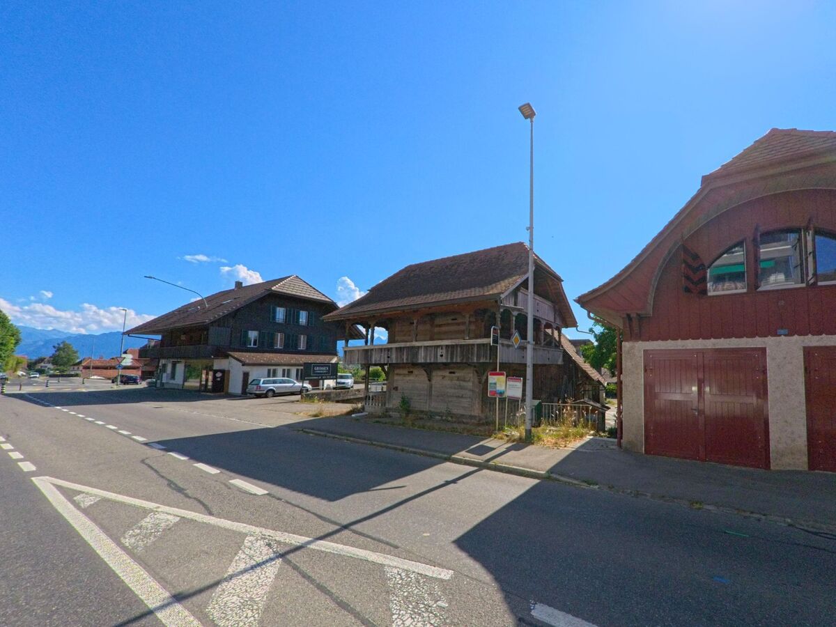 In Wichtrach (3114, Kanton Bern) stehen entlang einer breiten Strasse mehrere charakteristische Holzbauten, darunter ein Spycher, ein Geschäftshaus und ein rot gestrichenes Nebengebäude mit geschwungenem Dach.