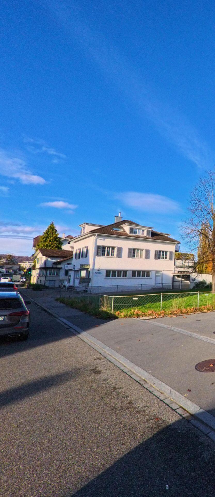 Ein helles Wohnhaus an einer Strassenecke mit Grünfläche und parkenden Autos liegt ruhig in Widnau (9443, Kanton St. Gallen) unter blauem Himmel.