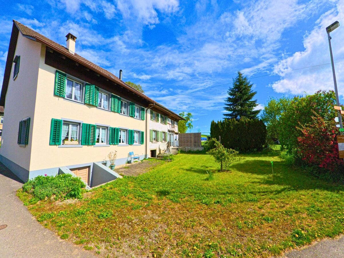 In Wiesendangen (8543, Kanton Zürich) zeigt das Bild ein gepflegtes Mehrfamilienhaus mit grünen Fensterläden, umgeben von einem kleinen Garten und Büschen.