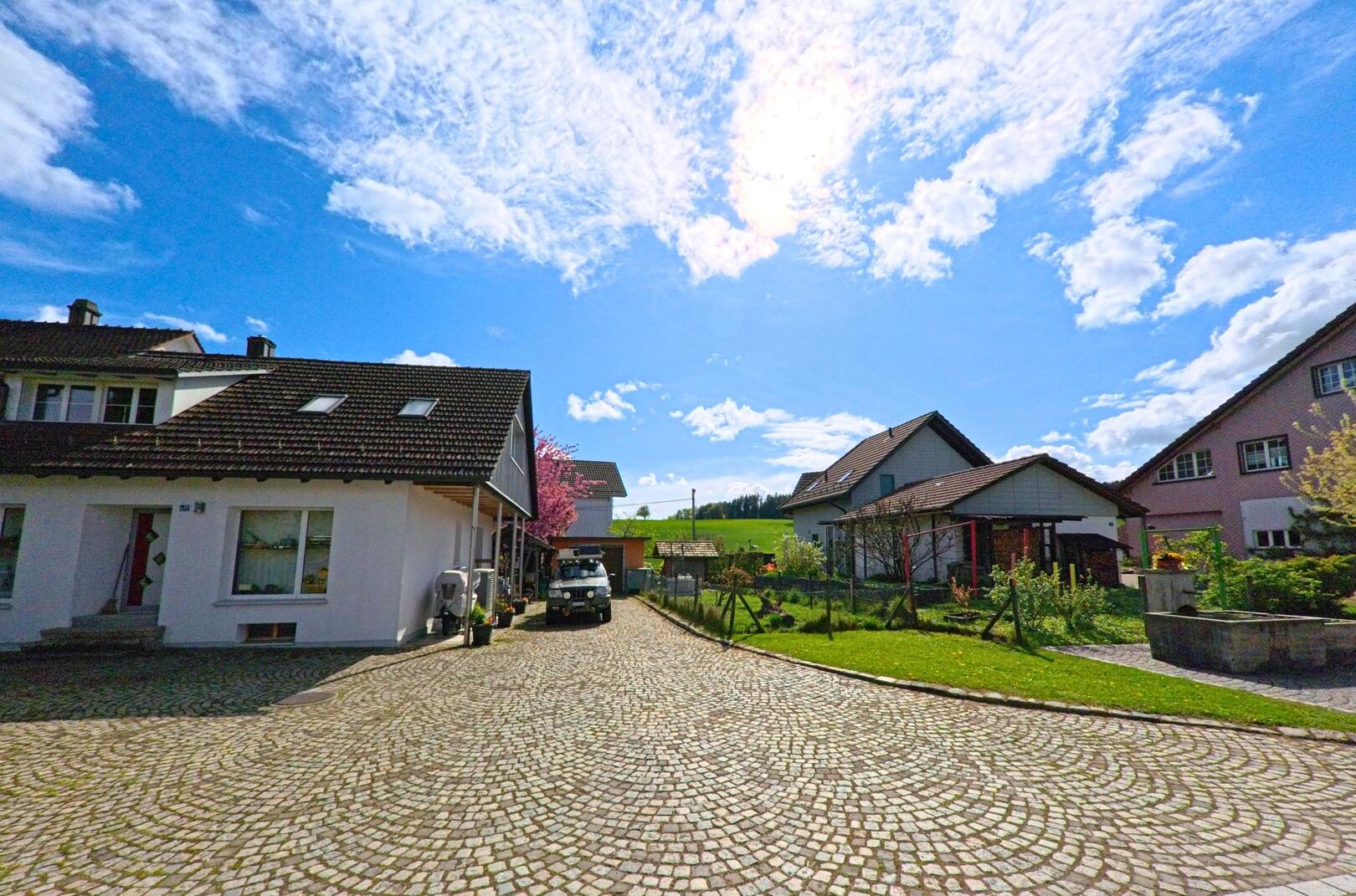 In Wiesendangen (8543, Kanton Zürich) führt ein gepflasterter Weg durch ein Wohnquartier mit gepflegten Einfamilienhäusern und kleinen Gärten unter Frühlingssonne.