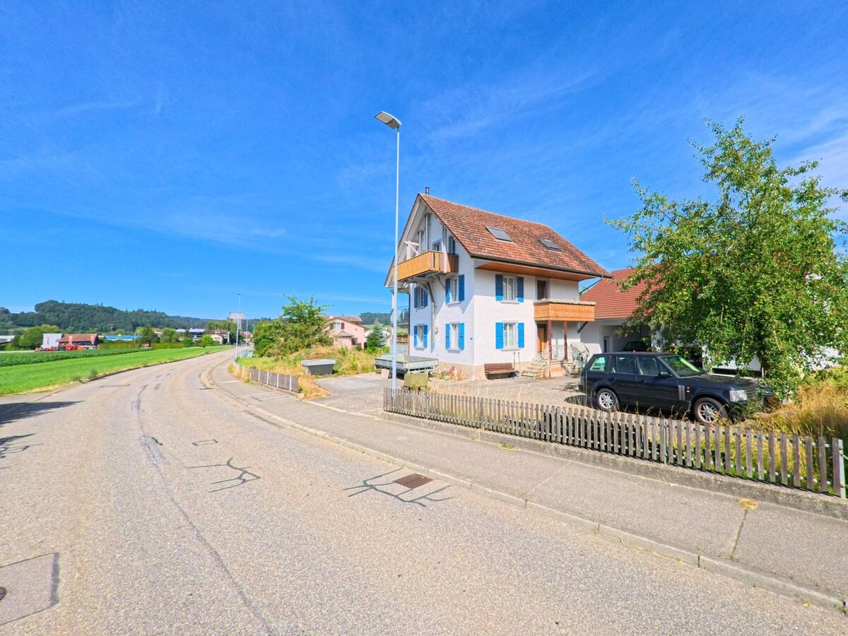 Ein hellblaues Einfamilienhaus mit Fensterlaeden steht am Rand einer ruhigen Strasse in Wikon (4806, Kanton Luzern), umgeben von Feldern und huegeliger Landschaft.