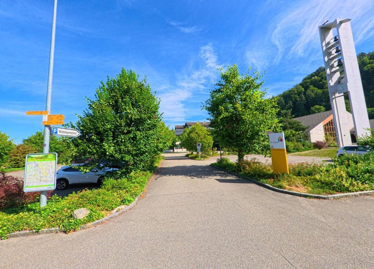 In Wikon (4806, Kanton Luzern) fuehrt ein Weg vorbei an einem modernen Kirchturm und Hinweisschildern zum Waldspielplatz, eingerahmt von Baeumen und Gruenanlagen.