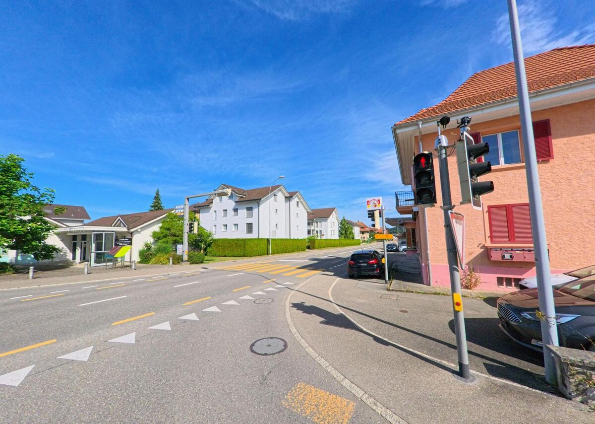 Eine Ampelanlage regelt den Verkehr an einer Kreuzung in Wikon (4806, Kanton Luzern), wo sich Wohnhaeuser, ein Wegweiser und ein rosafarbenes Gebaeude gegenueberstehen.