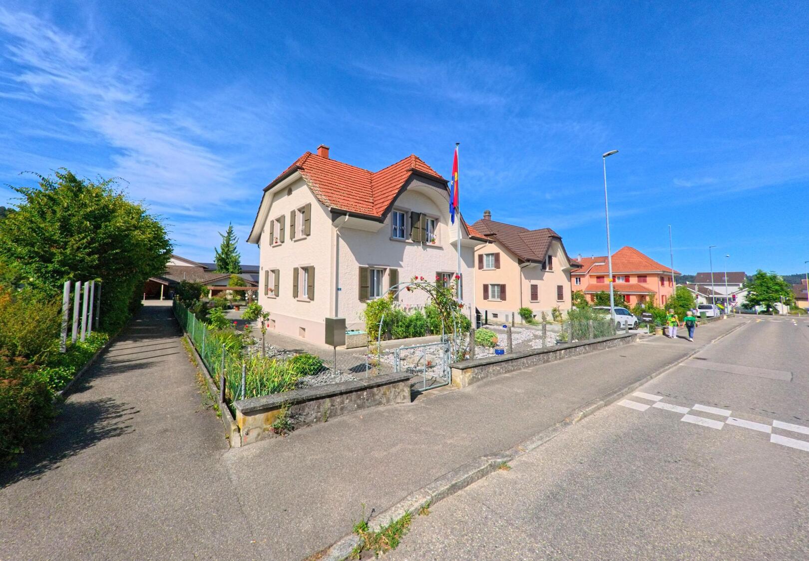 In der Gemeinde Wikon (4806, Kanton Luzern) säumen Einfamilienhäuser mit gepflegten Vorgärten und Schweizer Fahne eine ruhige Wohnstrasse unter blauem Himmel.